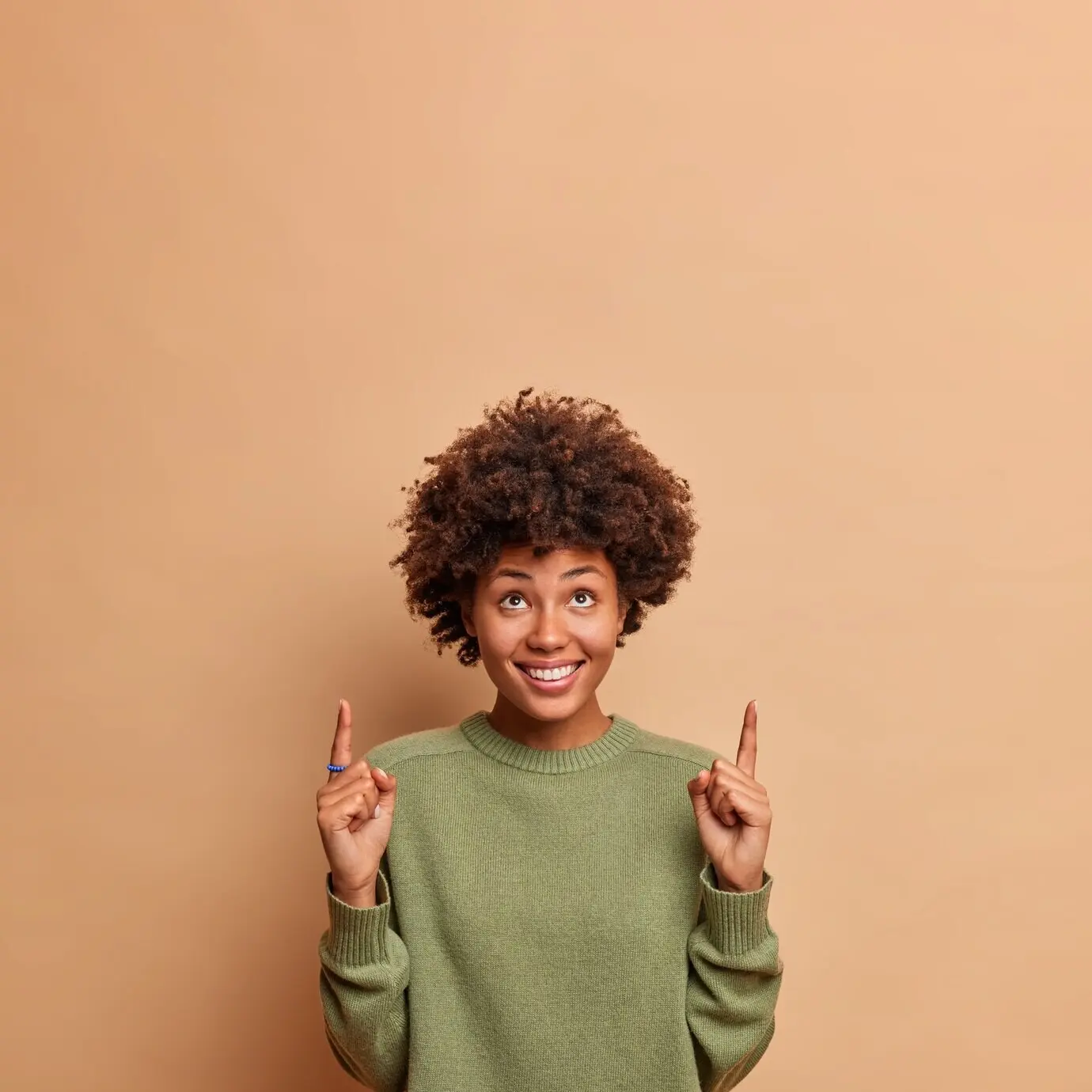 Vertical shot of a cheerful, joyful female model pointing upward with a broad smile, pleased to show copy space for your advertising content, wearing a casual jumper, isolated against a beige wall.