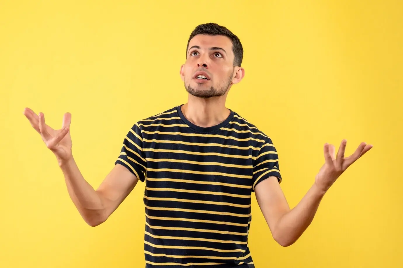 Front view of a handsome man in a black-and-white striped T-shirt looking up at the ceiling on an isolated yellow background.