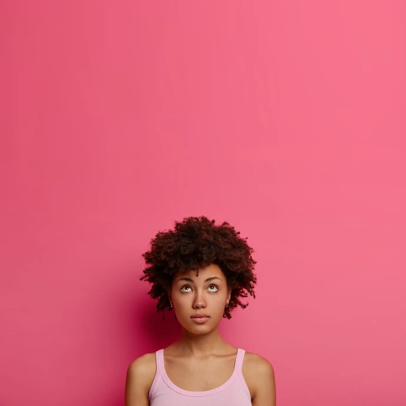 An African American woman with natural curly hair looks thoughtfully upward, has a thought in mind, wears casual clothes, stands against a pink wall with copy space for your promotion, and recalls something.
